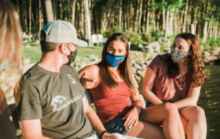Four people sit outdoors near water, wearing casual summer clothes and masks, engaging in conversation. Trees and rocks are visible in the background. Saint Joseph's College of Maine