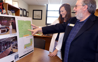 Steering A New Course 2 Two people standing by a table, pointing at architectural plans on a large board. They appear to be steering the discussion towards new design features shown on the display. Saint Joseph's College of Maine