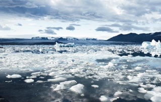 Opening The Arctic 5 A vast icy landscape with floating ice chunks on a still body of water, mountainous terrain in the background, and a partly cloudy sky above beckons an Arctic exploration. Saint Joseph's College of Maine