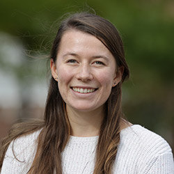 A woman with long brown hair wearing a white sweater is smiling while standing outdoors with a blurred green background, embodying the joy and dedication typical of someone pursuing a Bachelor of Science in Elementary Education. Saint Joseph's College of Maine