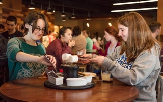 Tradition Of Hospitality 4 Two women prepare food at a round table in a busy, well-lit dining area, embodying the spirit of tradition. One scoops an ingredient while the other holds a plate, showcasing genuine hospitality. Several people are seated at other tables in the background. Saint Joseph's College of Maine