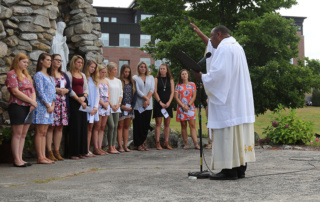 Following In The Footsteps Of Catherine Mcauley 3 A clergy member in white robes conducts an outdoor ceremony in front of a group of people, some holding papers, standing in front of a stone structure and greenery, following the footsteps of Catherine McAuley. Saint Joseph's College of Maine