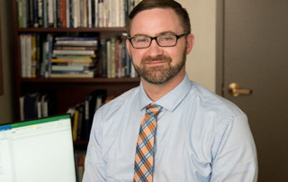 A Commitment To Justice 7 A man with short hair and glasses, dressed in a light blue shirt and orange plaid tie, sits in an office beside a computer and bookshelves, surrounded by legal texts that signify his commitment to justice. Saint Joseph's College of Maine
