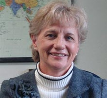 Online Radiology Program Grows 7 A woman with short-blonde hair smiles, wearing a grey blazer over a white turtleneck. Behind her, a world map hints at the global reach of the online radiology program she's discussing. Saint Joseph's College of Maine