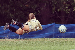 Brooke Troup Talks About The New Track And Field Facility 8 Soccer player Brooke Troup in mid-air dive attempting to reach a ball on a grassy field with blue netting in the background at the track and field facility. Saint Joseph's College of Maine