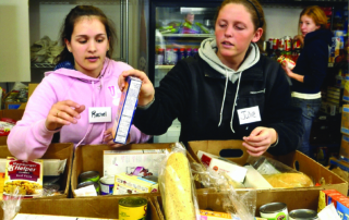 Reflections On A Cupboard That’s Never Empty 7 Two women, wearing name tags reading "Rachel" and "Julie", sort food items in boxes at a food bank. Reflections of their commitment glimmer as they work diligently. Another person in the background also handles food items, ensuring no cupboard is ever empty. Saint Joseph's College of Maine