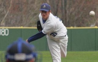 Former Monks Power Pitcher Shows Major Moves 4 A power pitcher in a white and blue uniform throws a pitch towards the batter on a grass field, with the catcher seen in the foreground. Saint Joseph's College of Maine