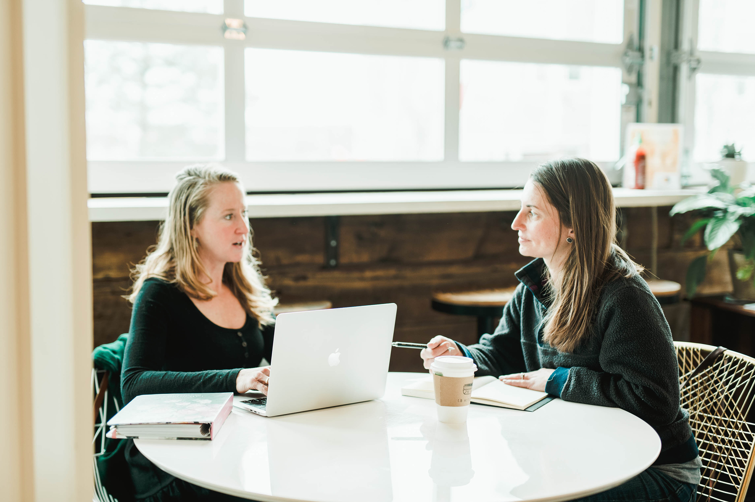 Two women are sitting at a round table with a laptop, a notebook, and a coffee cup, engaged in a conversation about online health administration programs. Saint Joseph's College of Maine