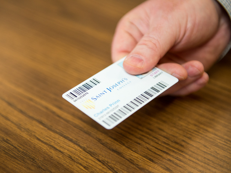 A hand holding a Saint Joseph's College ID card with barcodes and the name Charles Primn visible on the wooden table background, right next to an entrance brochure for Wellehan Library. Saint Joseph's College of Maine