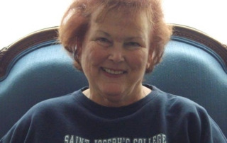 Meet Cornelia Baker '85 1 Cornelia Baker, with her short, curly red hair, sits in a blue upholstered chair, smiling. She is wearing a navy sweatshirt that proudly reads "Saint Joseph's College of Maine Alumni. Saint Joseph's College of Maine