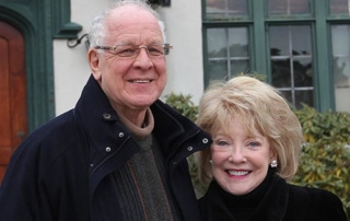 Bridging The Distance 5 An older man in a black coat and an older woman in a black coat stand smiling in front of a house with green trim, symbolizing their way of bridging the distance. Saint Joseph's College of Maine
