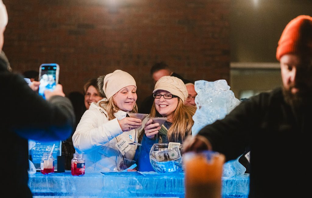 Alumni 5 Two women in winter hats and coats clink drinks and smile at an ice bar in Maine, with other people around them. One person in the foreground holds a drink beside tip jars and ice sculptures on the bar, capturing classic college night vibes. Saint Joseph's College of Maine