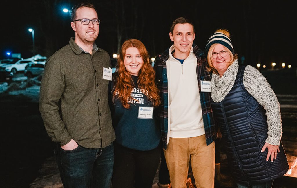 Alumni 6 Four people stand close together and smile at the camera outdoors at night in Maine. Dressed warmly in winter clothing, they pose with snow on the ground, with lights and cars twinkling in the background. Saint Joseph's College of Maine