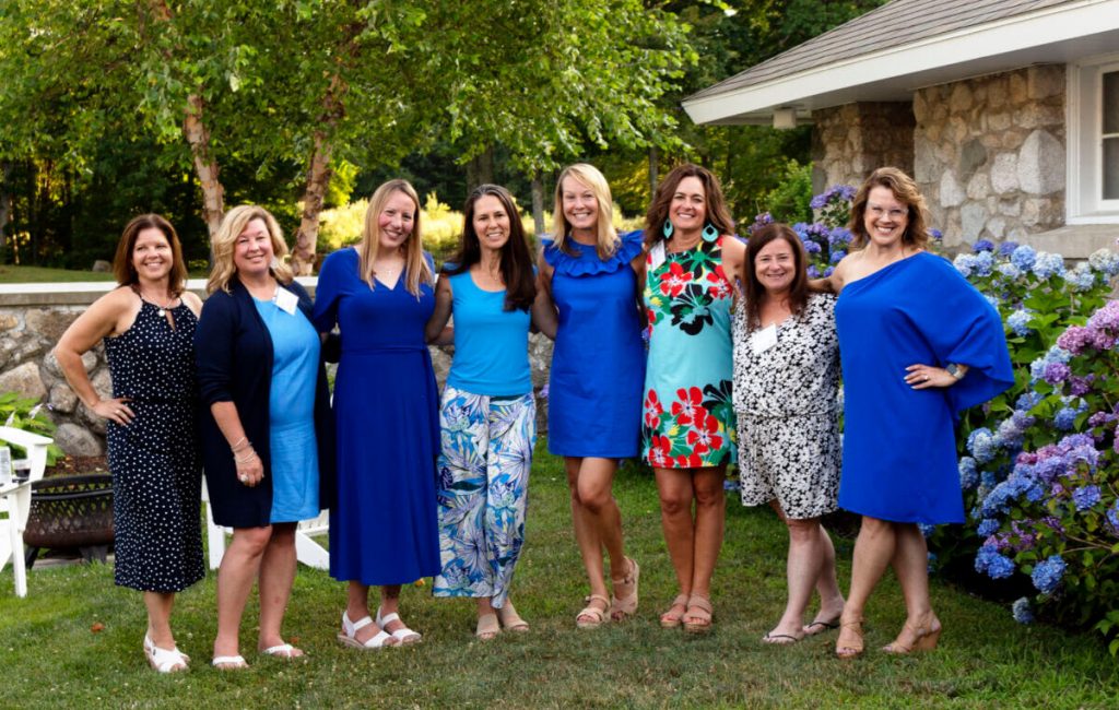 Eight women stand together on the grass in Maine, smiling at the camera. They are dressed in blue and patterned summer dresses. Green trees, a stone house, and blooming hydrangeas are visible in the background. It’s a perfect college reunion. Saint Joseph's College of Maine