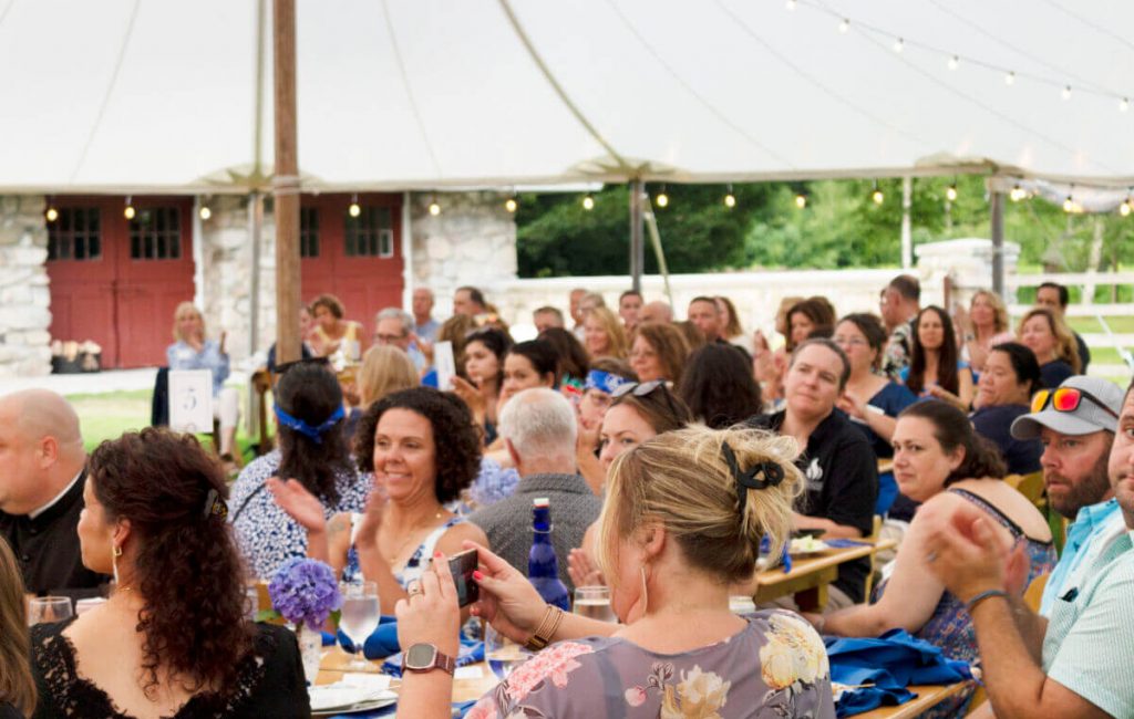 A large group of people sits at picnic tables under a tent, clapping and smiling. String lights hang overhead, giving the festive outdoor setting—reminiscent of a lively college gathering in Maine—a warm and inviting glow amid rustic buildings. Saint Joseph's College of Maine
