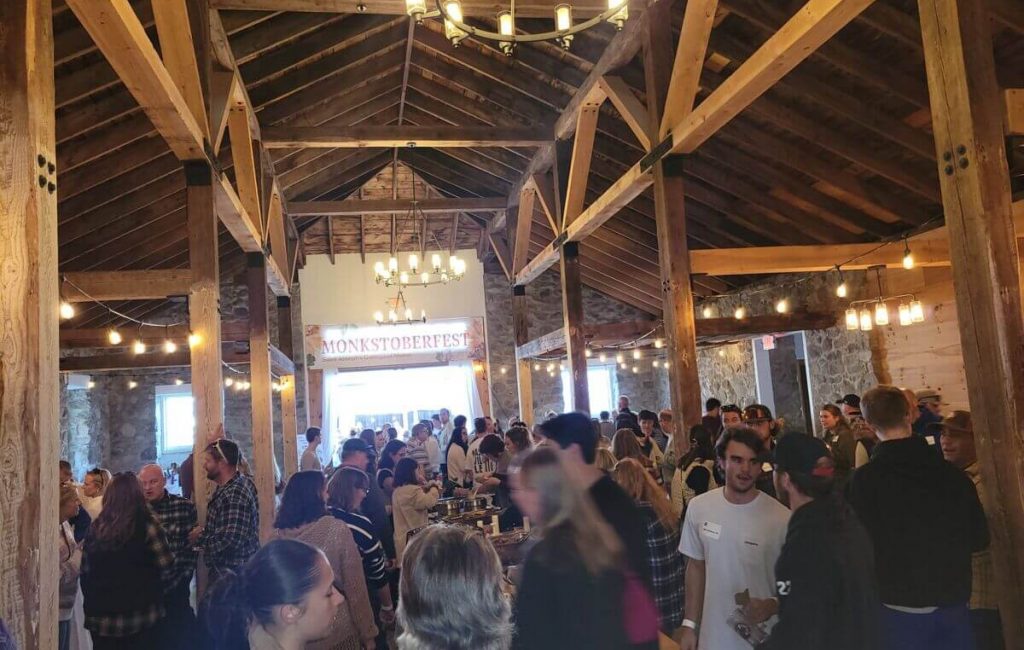 Alumni 4 A large group of people, including local college students, gathers inside a rustic wooden hall with exposed beams and chandeliers, attending an event called Monkstoberfest, as seen on a banner in the background. Saint Joseph's College of Maine
