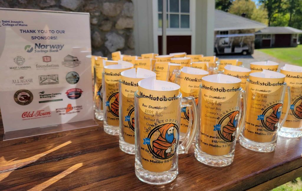 Alumni 3 Several beer glasses with colorful Oktoberfest designs are arranged on a wooden table outdoors at a Maine college event. A sponsor sign with various logos is displayed next to the glasses as sunlight casts bright shadows on the scene. Saint Joseph's College of Maine