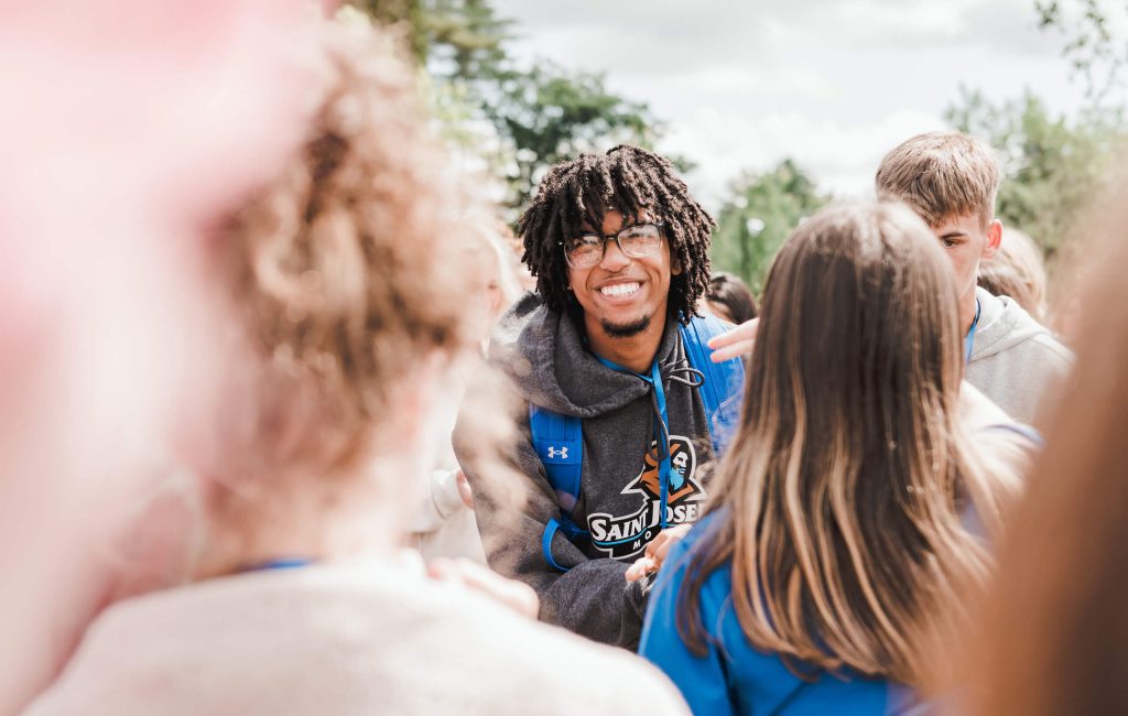 A person with dreadlocks and glasses smiles while standing in the middle of a group of people outdoors on a sunny day, capturing the essence of student life. Saint Joseph's College of Maine