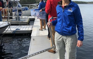 New Sebago Lake Buoy Collects Real-Time Water Quality Data For Scientists, Students, And The Public 7 A person wearing a blue jacket and sunglasses stands on a dock beside Sebago Lake, smiling and holding a tool. Several other people, including students and scientists, are on the dock, collecting real-time water quality data while boats are docked nearby. Saint Joseph's College of Maine