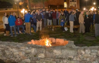 A Welcomed Light 5 A group of people gather around an outdoor fire pit at night, with some seated on benches and others standing. The warm glow acts as a welcomed light, while nearby buildings and streetlights provide additional lighting, creating a cozy ambiance that feels like an extension of thoughtful home decor. Saint Joseph's College of Maine