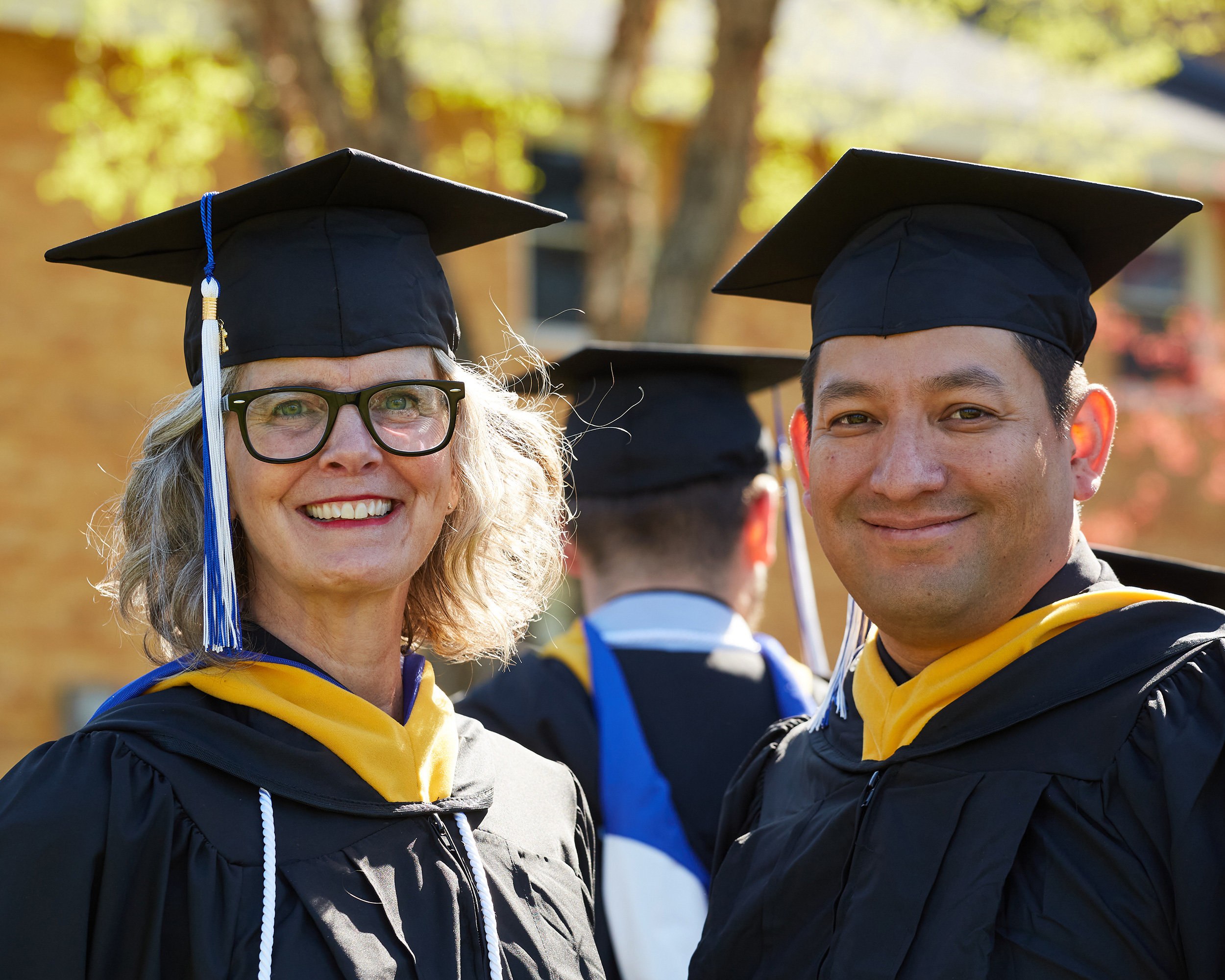 Two graduates in caps and gowns smile at the camera during an outdoor graduation ceremony, celebrating their journey from admissions to achievement. Saint Joseph's College of Maine