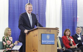 Harold Alfond Foundation Gives $1.5 Million Challenge Grant For Saint Joseph’s College’s Center For Nursing Excellence 3 A man speaks passionately at a podium labeled "Saint Joseph's College," while five seated individuals listen attentively against a backdrop of blue curtains, highlighting the significance of the Harold Alfond Foundation Challenge Grant to the institution. Saint Joseph's College of Maine