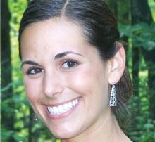 Double Alumni 9 A double alumni with dark hair tied back, wearing earrings and a black top, smiles at the camera with a forested area in the background. Saint Joseph's College of Maine