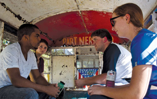 Without Warning 3 Four people are sitting inside a worn-out, partially painted vehicle, engaged in conversation. Without warning, one person reveals a green object while another clutches a water bottle. Saint Joseph's College of Maine