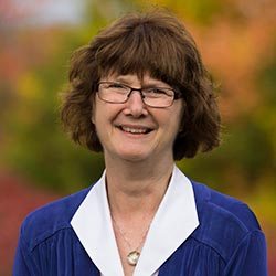 Giving 11 A woman with short brown hair and glasses is smiling. She is wearing a blue jacket over a white collared shirt. The background is blurred with hints of autumn colors, reminiscent of the beautiful campus for those interested in ways to give back to Saint Joseph's College of Maine. Saint Joseph's College of Maine