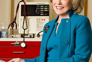 Saint Joseph'S College Receives Largest Individual Gift In Its History From Dr. Jeanne Donlevy Arnold For Center For Nursing Innovation 7 A smiling woman in a teal suit holds a clipboard in a medical office at Saint Joseph's College, with a defibrillator and medical equipment in the background. Saint Joseph's College of Maine