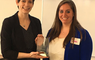 College And Jenna Chase Recognized By Breathe Easy Coalition Of Maine For Gold Star Standard Of Excellence 3 Two women are standing indoors. One, Jenna Chase, holds an award while the other stands beside her, smiling. Both are dressed in business attire, representing the Breathe Easy Coalition during a college event. Saint Joseph's College of Maine