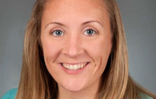 Light Bulb Moments 5 A woman with long blonde hair, wearing a teal top, smiles at the camera against a gray background, capturing one of those light bulb moments. Saint Joseph's College of Maine