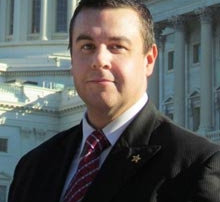 Whose Idea Is It, Anyway? 2 A man in a black suit and red tie, whose confident demeanor catches the eye, stands in front of a large classical building with columns on a sunny day. Saint Joseph's College of Maine