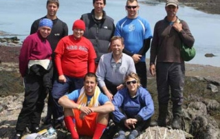 Out Of The Classroom, Into The Bay 7 A group of nine people wearing casual outdoor attire poses on rocky terrain by the bay. Some are standing while others are seated. The background features a shoreline and scattered rocks, creating an informal outdoor classroom setting. Saint Joseph's College of Maine