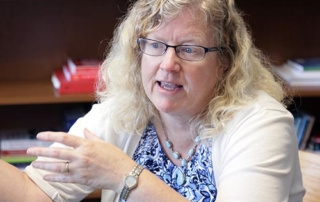 Bsw Opens Many Doors 7 A woman with curly blonde hair and glasses is speaking while gesturing with her hand. She is wearing a white cardigan and a printed top. Books are visible on a shelf in the background, suggesting that knowledge opens many doors to new opportunities. Saint Joseph's College of Maine