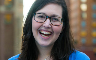Commencement To Feature Award-Winning Catholic Journalist/Author Kerry Weber As Speaker And One Of Three Honorary Degree Recipients 6 Kerry Weber, a Catholic journalist with long dark hair and glasses, is seen smiling in a blue cardigan and necklace, standing in front of a blurred urban background. Saint Joseph's College of Maine