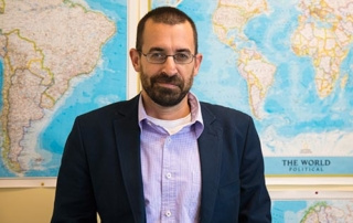 Beyond Partisanship 1 A man with glasses and a beard, dressed in a blazer and a collared shirt, stands in front of a wall adorned with world maps, exuding an air of intellect that transcends beyond partisanship. Saint Joseph's College of Maine