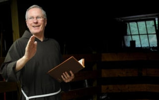 New Chaplain Joins Mercy Center Staff 6 A man in a brown robe with a white cord, likely a chaplain from the Mercy Center, holds an open book and gestures with his hand, standing in a dimly lit room with a window in the background. Saint Joseph's College of Maine