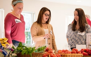 Work With Purpose 4 Three women stand at a table with assorted fresh produce, engaged with purpose. Two have name tags reading "Maria" and "Emily." One woman is selecting a tomato, focused on her work. Saint Joseph's College of Maine