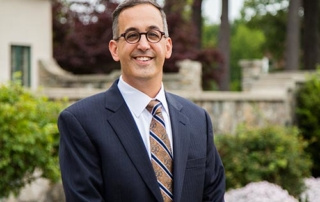 Catherine’s Legacy 3 A smiling man in a suit and tie stands outdoors in front of a garden with greenery and stone structures, embodying Catherine’s Legacy. Saint Joseph's College of Maine
