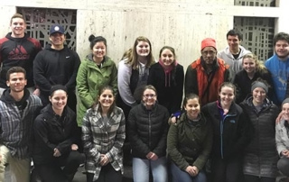 Students And Staff Participate In Midnight Run 3 A group of 18 students, wearing winter clothing, pose together indoors in front of a plain background, preparing for the Midnight Run. Saint Joseph's College of Maine