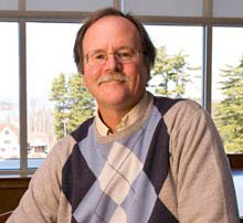 New books by faculty 3 A man with glasses and a mustache is wearing a sweater with a diamond pattern, sitting indoors in front of a window with trees outside, reading new books. Saint Joseph's College of Maine
