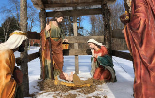 Outdoor Nativity scene in the snow, featuring statues of Mary, Joseph, baby Jesus in a manger, and two other figures kneeling. Saint Joseph's College of Maine
