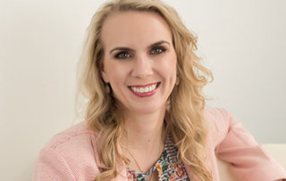 April Chase – Earning Msn, Delivering The Online College Graduates Speech At May 2018 Commencement 10 A woman with long, wavy blonde hair is smiling, wearing a light pink blazer and a floral-patterned top, sitting against a light-colored background, ready to deliver her commencement speech for an online college. Saint Joseph's College of Maine