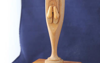 Statue Story 6 A wooden statue titled "The Original Monk," set against a blue background, sits on a wooden base with a gold plaque reading, "Donated by Dr. Christopher M. Kiernan." This statue tells the story of art preserved through time. Saint Joseph's College of Maine