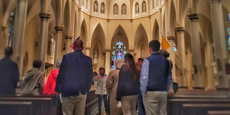 Overview-Center-Faith-Spirituality A group of people stands inside a large church with high arches and stained glass windows, facing a guide from the Center for Faith and Spirituality who is speaking in front of them. Saint Joseph's College of Maine
