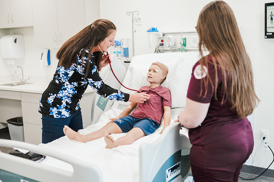 Partners5 A healthcare provider, demonstrating the value of partnerships in patient care, uses a stethoscope to listen to the chest of a young patient sitting on a hospital bed while another healthcare worker stands nearby. Saint Joseph's College of Maine