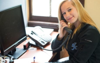 Fall Phonathon Reaches Thousands 3 A woman with long blonde hair is sitting at her desk, talking on the phone, and writing on paper during the Fall Phonathon. A computer monitor and telephone are also on the desk. She is smiling brightly and looking at the camera, ready to connect with thousands of potential donors. Saint Joseph's College of Maine