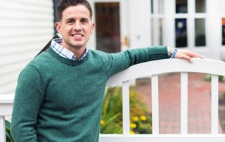 Giving Back 1 A person wearing a green sweater and checkered shirt stands outdoors, leaning on a white fence with a building and greenery in the background, embodying the spirit of giving back. Saint Joseph's College of Maine