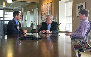 Curating Governor Brennan’s Legacy 10 Three men in business attire are seated at a large conference table having a conversation in an office setting, discussing Governor Brennan's legacy. Saint Joseph's College of Maine